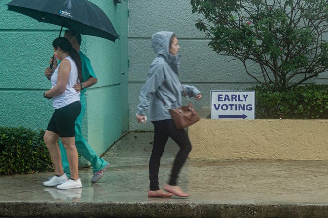 A couple walks back to their car under their umbrella having just voted early, as a woman in a rain slicker walks toward the line for early voting at the Palm Beach County Library on Summit Blvd., Monday, October 19, 2020. Statewide early voting started today. (JOSEPH FORZANO / THE PALM BEACH POST)