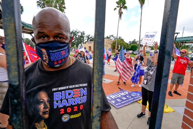 James Dozier, front left, a supporter of Democratic presidential candidate former Vice President Joe Biden listens to former President Barack Obama speak at a rally as President Donald Trump supporters shout and wave flags behind him Tuesday, Oct. 27, 2020, in Orlando, Fla.